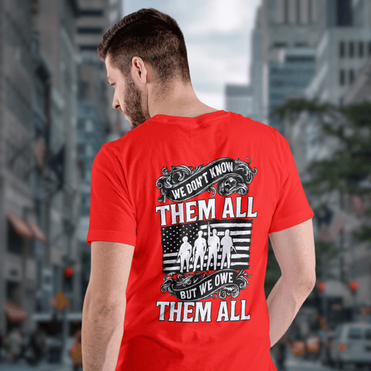 Man wearing a red patriotic t-shirt with a military tribute design on the back that reads 'We Don’t Know Them All, But We Owe Them All' in bold red and white text over a distressed American flag and soldier silhouettes, standing in a city street background.