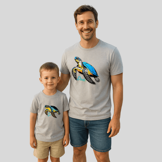 Father and son wearing matching gray t-shirts with colorful sea turtle designs labeled “Dude” and “Lil Dude,” standing together and smiling.